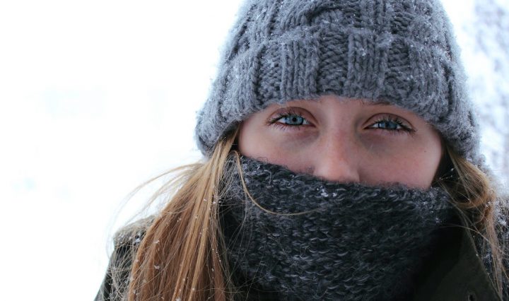 A Woman With Blue Eyes Covering Her Face And Head With A Grey Hat And Scarf